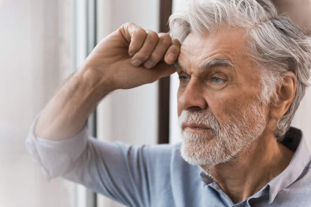 Ein älterer Mann lehnt mit seinem Ellenbogen und verzweifeltem Blick an einer Fensterscheibe.