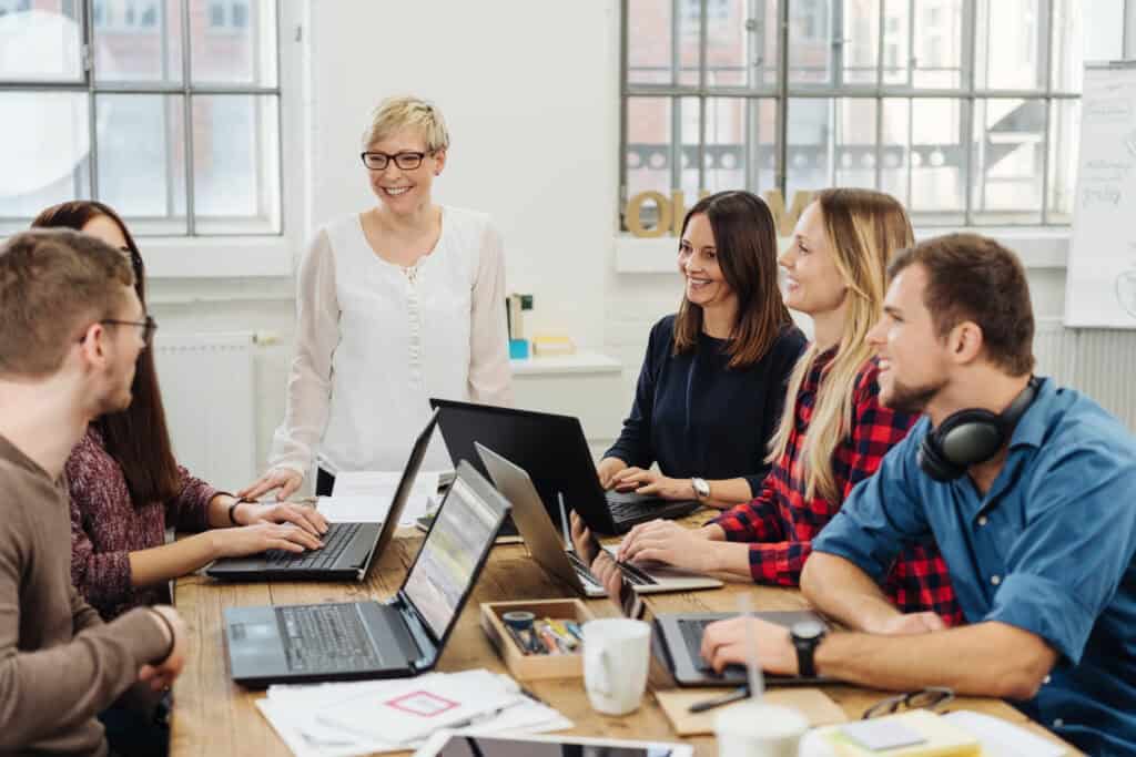 Eine Frau spricht stehend vor ihren Arbeitskollegen, die alle gemeinsam an einem Tisch vor ihren Laptops sitzen, ihr zuhören und sich austauschen.