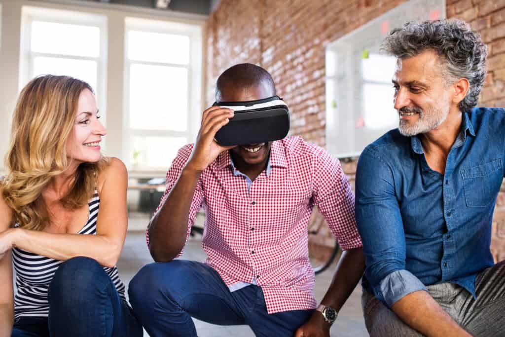 Ein Mann mit rotem Hemd probiert staunend eine VR-Brille aus. Ein anderer Mann und eine Frau, neben ihm hockend, sehen ihm glücklich dabei zu.