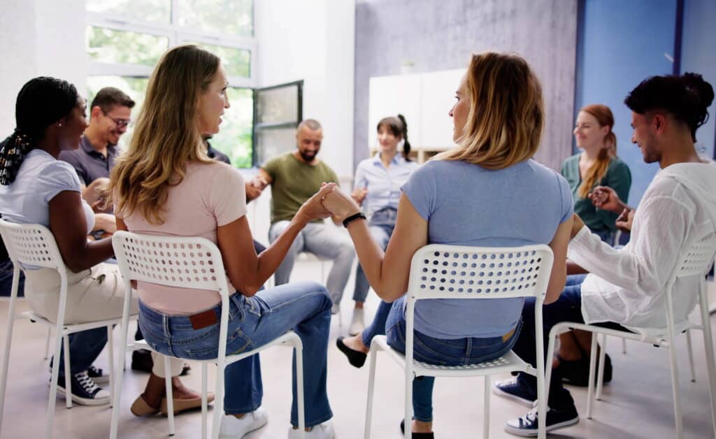 Eine Gruppe aus ca. 10 Menschen sitzt im Stuhlkreis und hält sich an den Händen.